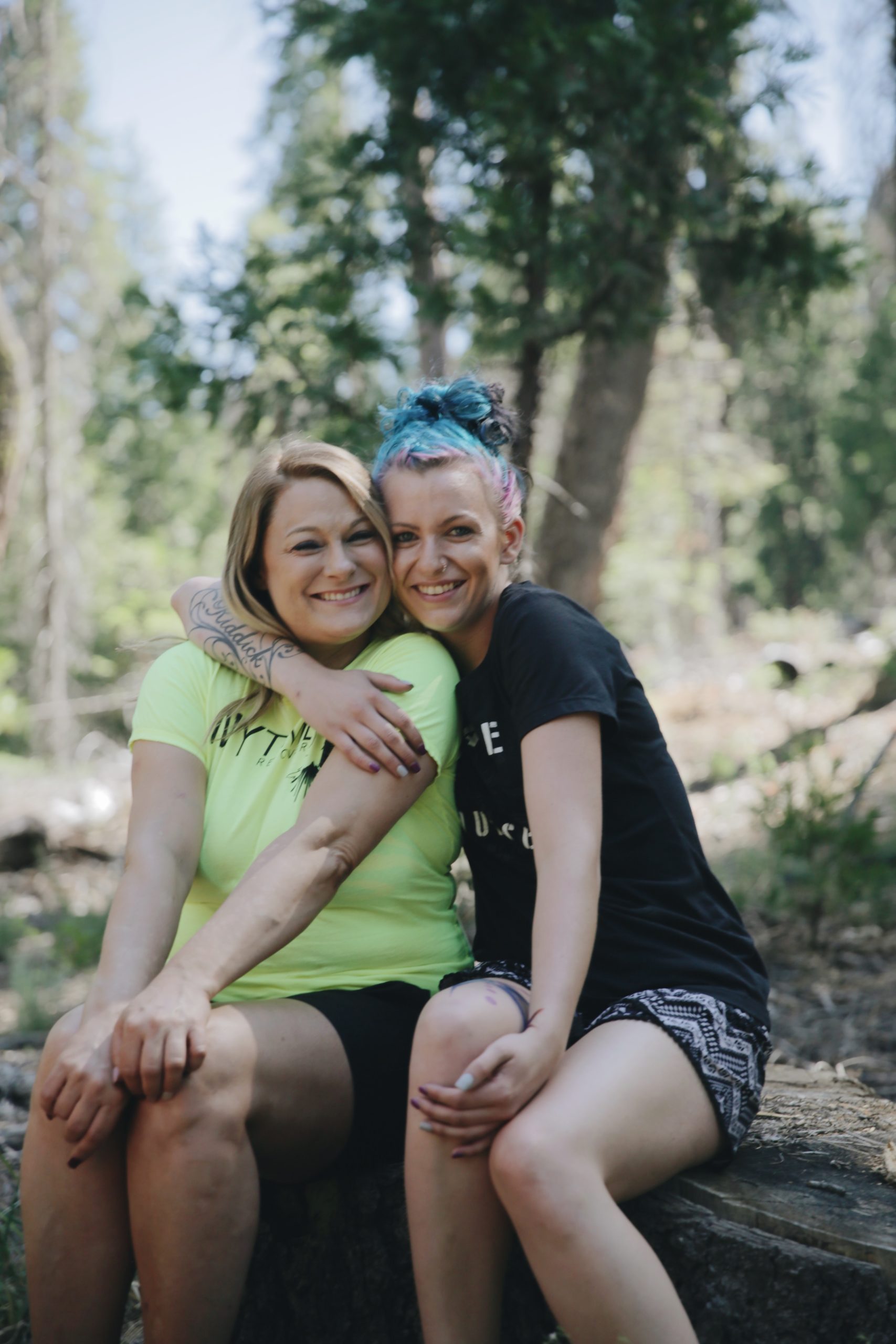 Two women sitting on a log in a forest setting, smiling and hugging each other during an outdoor adventure therapy session.